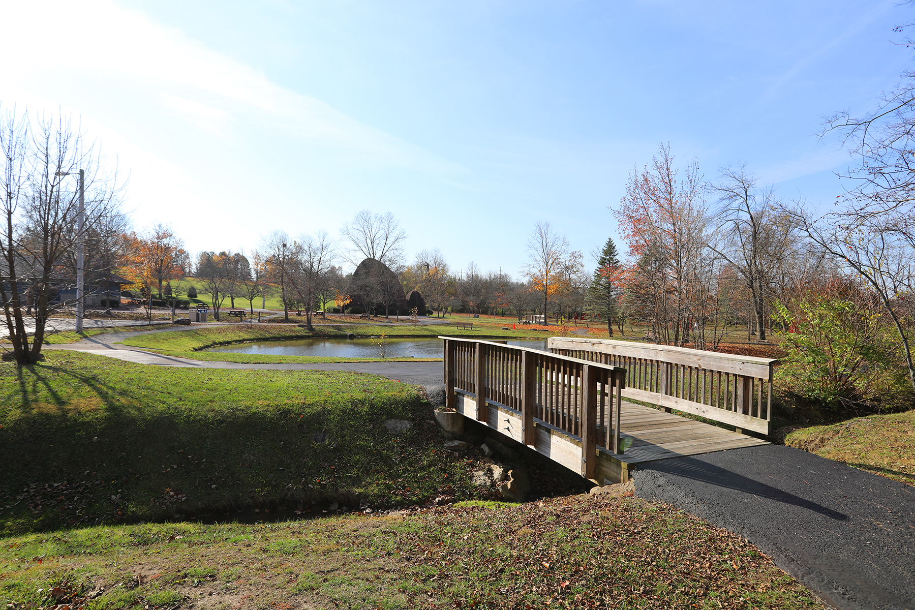 A large park in midday featuring a small bridge, a pond, and well-maintained landscaping.