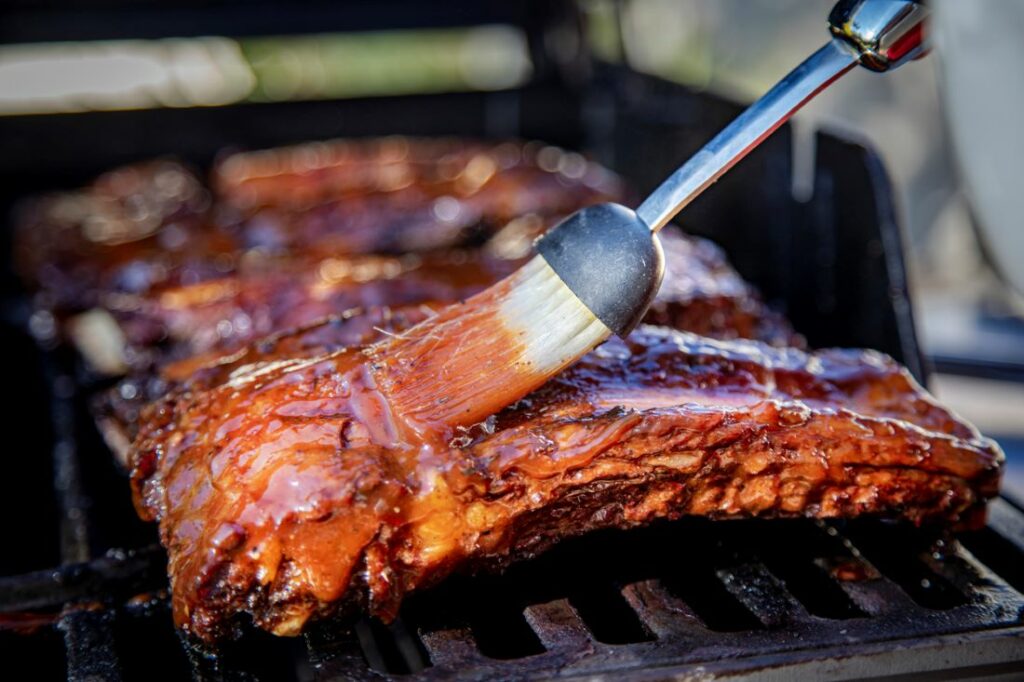 Beef ribs being basted with barbecue sauce on a grill with a basting brush