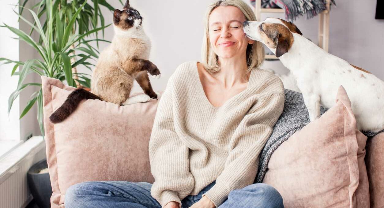 Woman sits on couch next to small dog and cat, smiling
