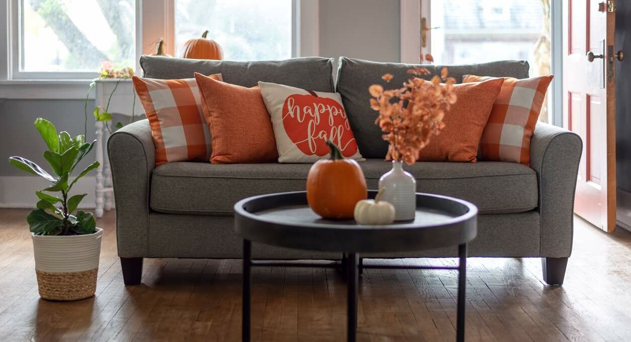 A living room decorated for autumn with orange pillows on a gray sofa and pumpkins on a black coffee table. A throw pillow reads “Happy Fall”