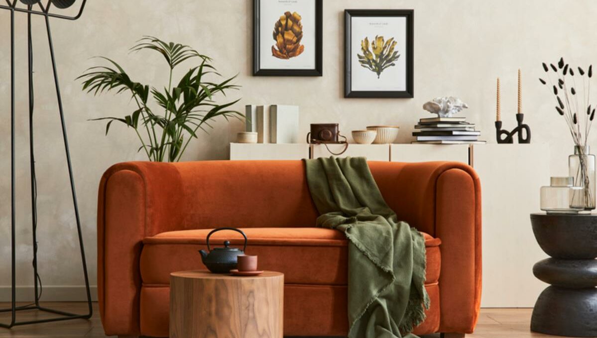 A decorated living room featuring an orange velvet couch, green throw blanket, natural art, a round wooden table, plants, books, and candles.