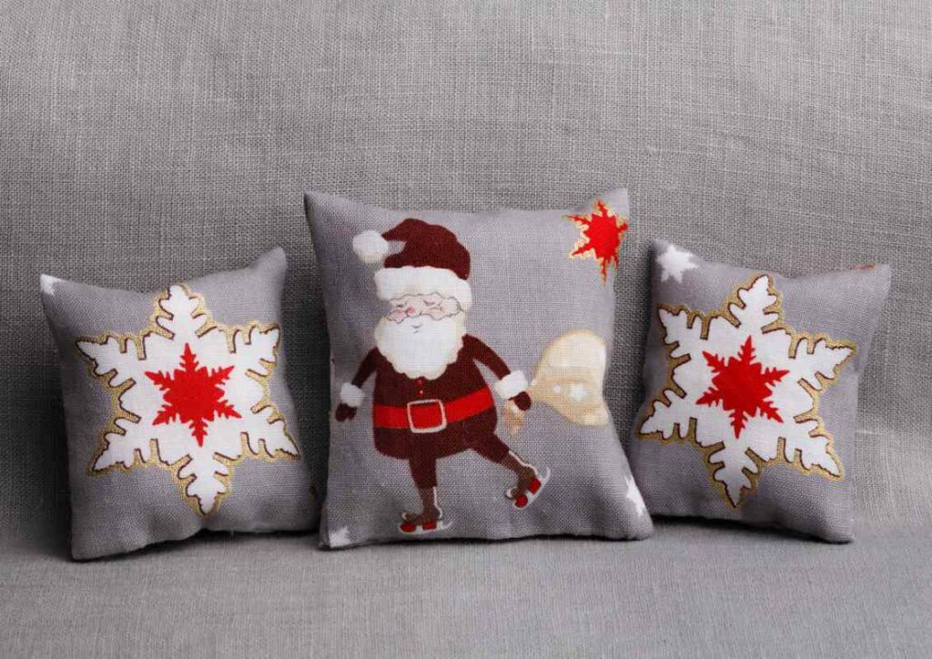 Christmas throw pillows with red and white snowflakes and Santa Claus designs displayed on a gray sofa.