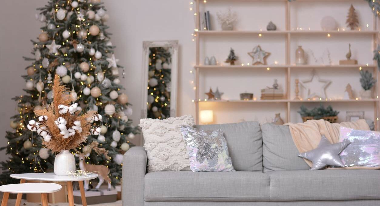 A modern apartment living room is decorated with silver and gold Christmas accents, with fairy lights, stars, and a Christmas tree in the background.