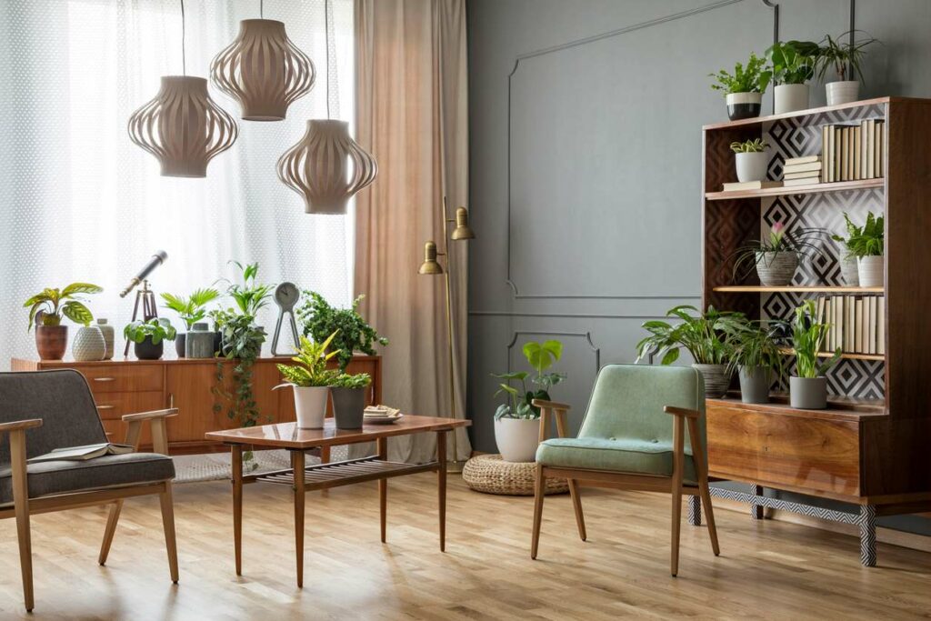 Wooden table with plants between green armchairs in a gray room with lamps and more plants.