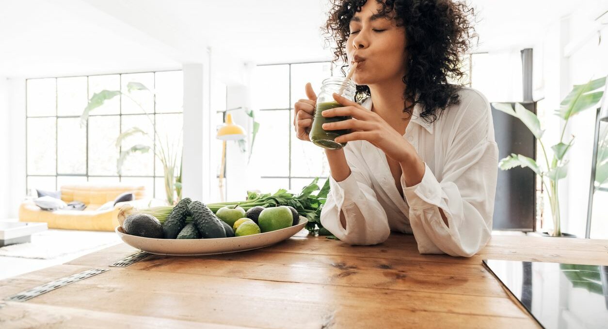 In a bright loft apartment, a young woman drinks green juice from a glass cup with a reusable bamboo straw.