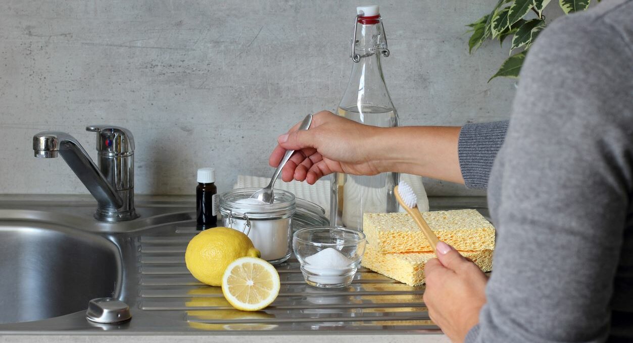 A view of someone making a DIY cleaner with baking soda, vinegar, a lemon, sponges, and a brush at the sink.