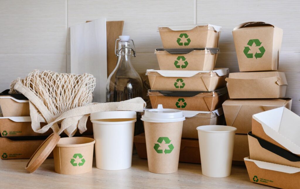 Food cartons with a green recycling symbol, a glass bottle, and a reusable shopping bag on a table.