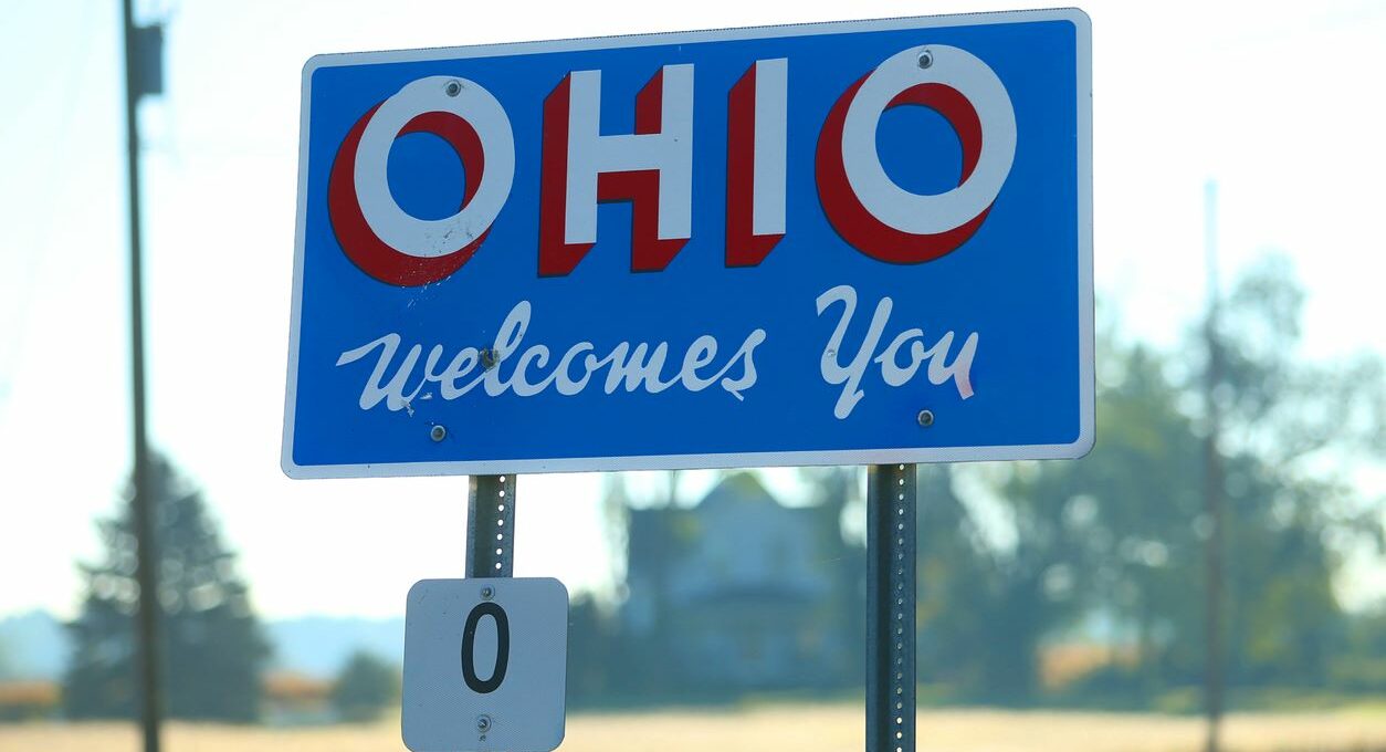 A blue sign reads “Ohio welcomes you” in white text with a blurred scenic background.