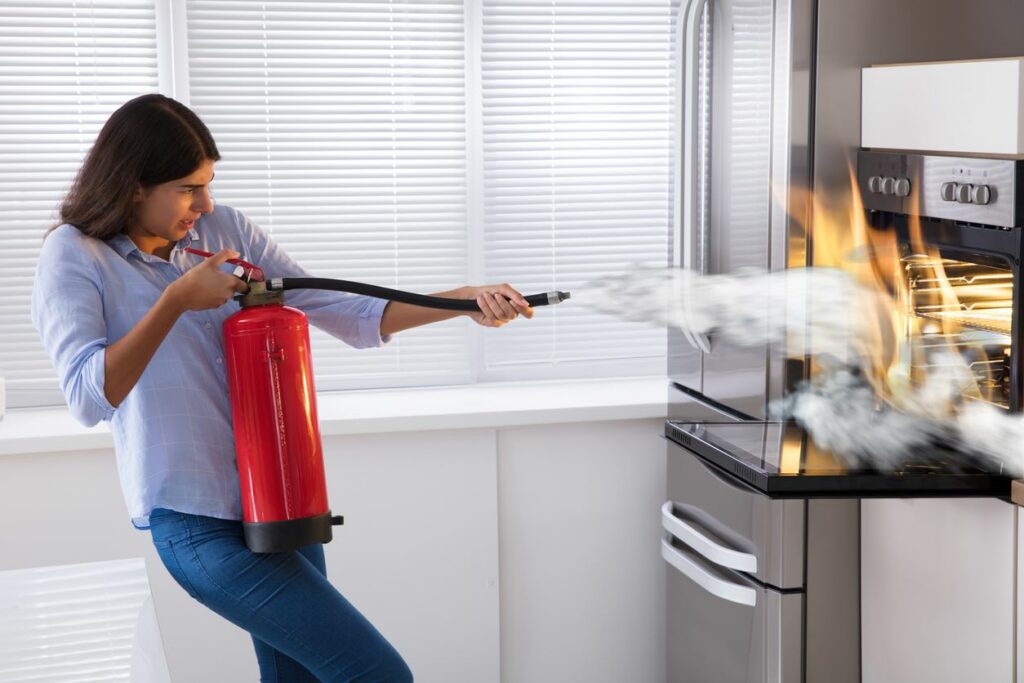 A young woman uses a fire extinguisher to snuff out an oven fire in the kitchen.