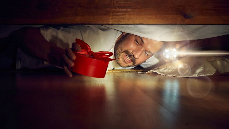 A man holding a flashlight places a red present with a bow under the bed.