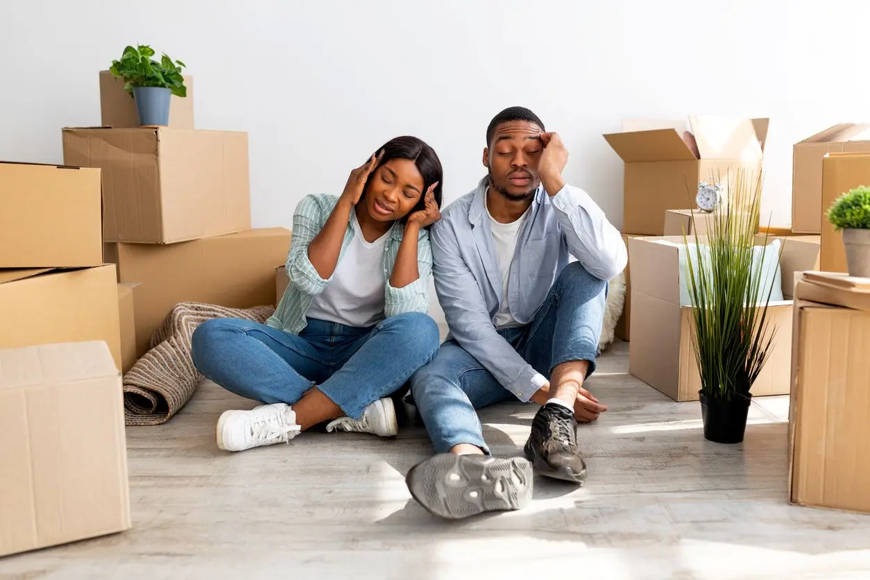 An exhausted couple sits together on the floor, surrounded by moving boxes and three houseplants.