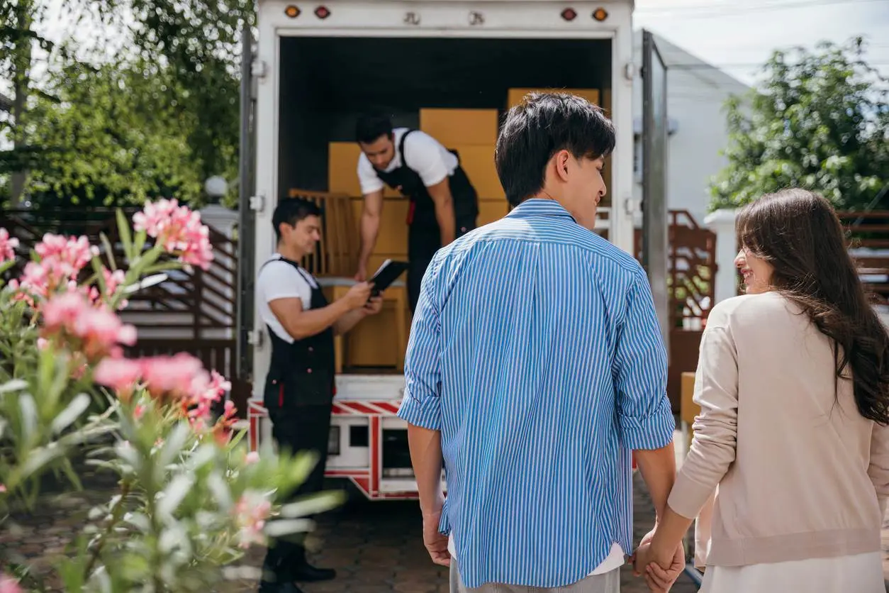 A smiling couple holds hands as professional movers check all the boxes in the truck.