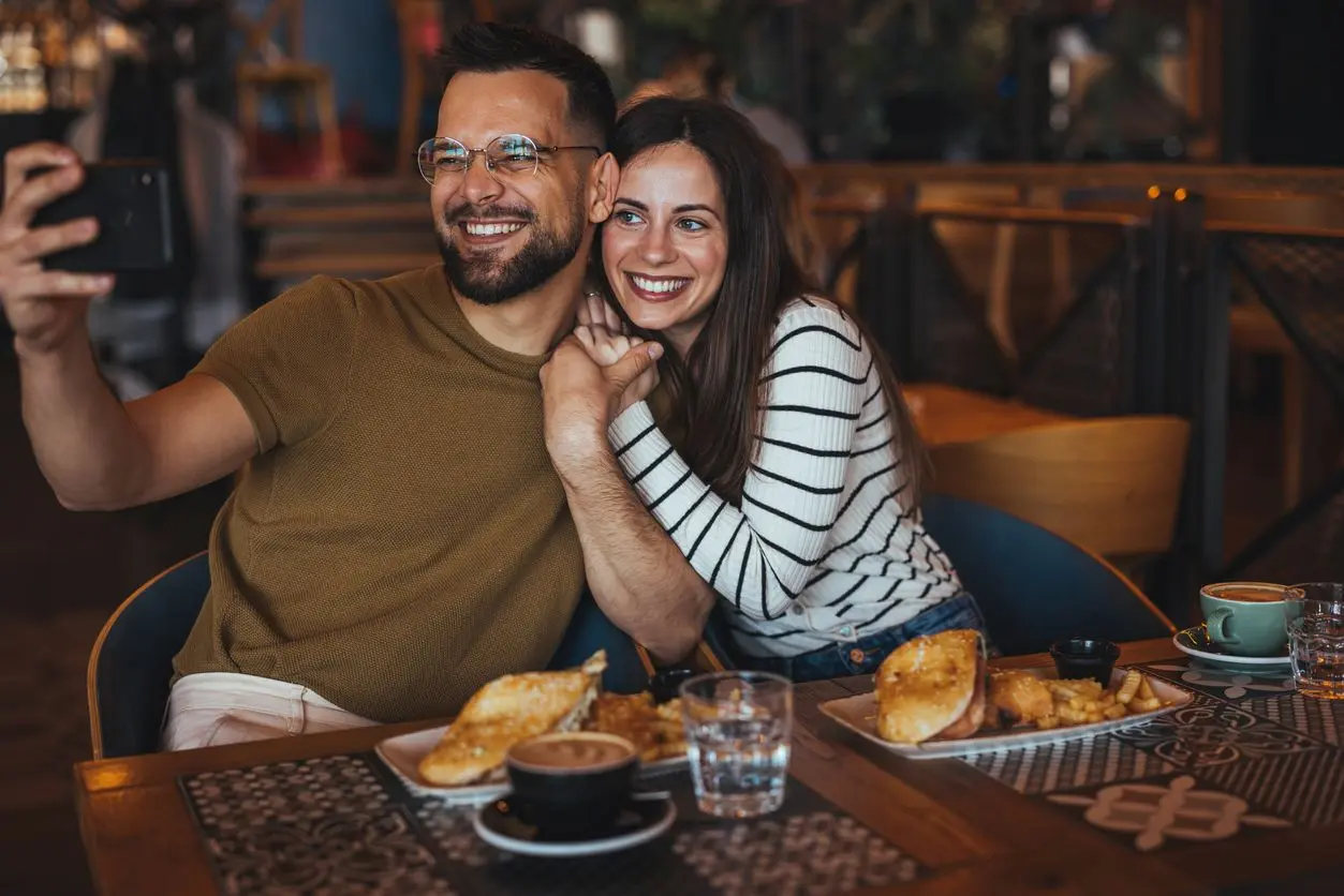 A smiling couple at a restaurant pose for a selfie with food on the table.