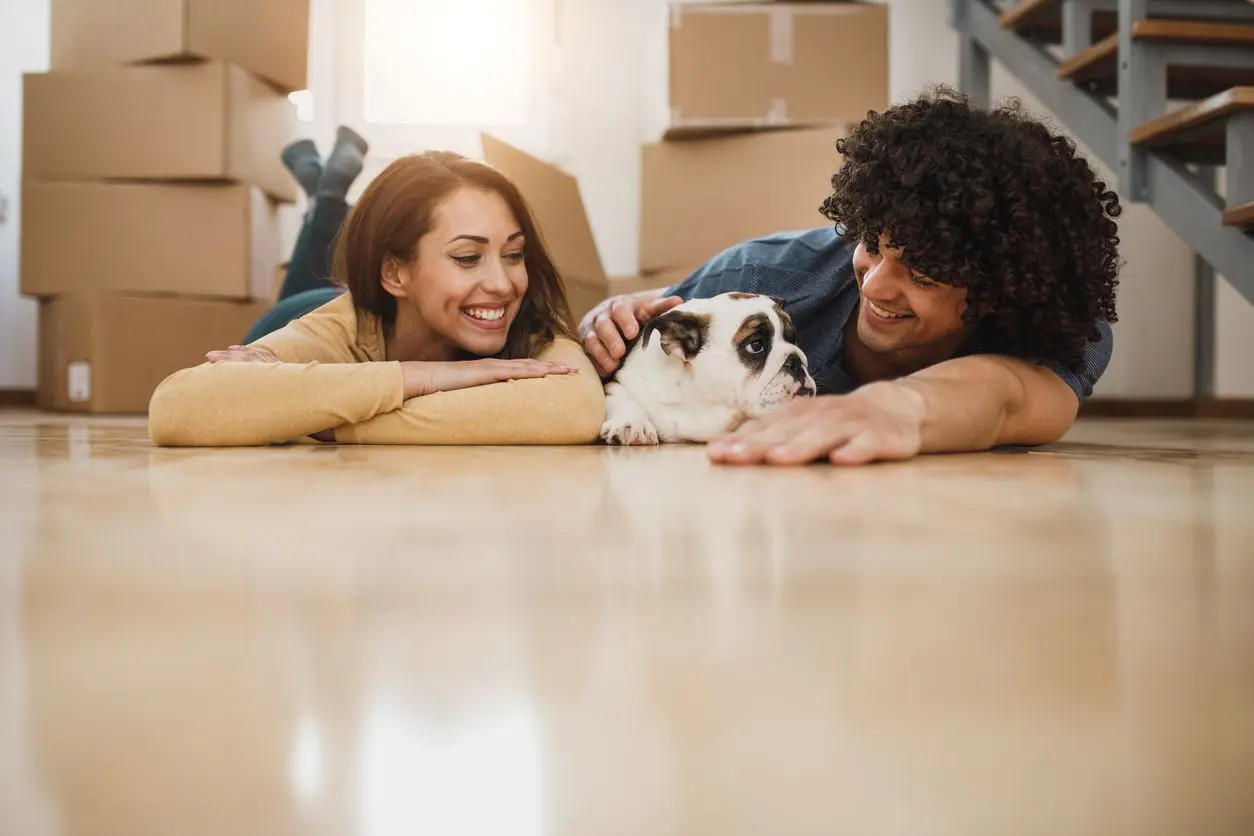 A smiling young couple lay on the floor with a bulldog between them, surrounded by moving boxes.