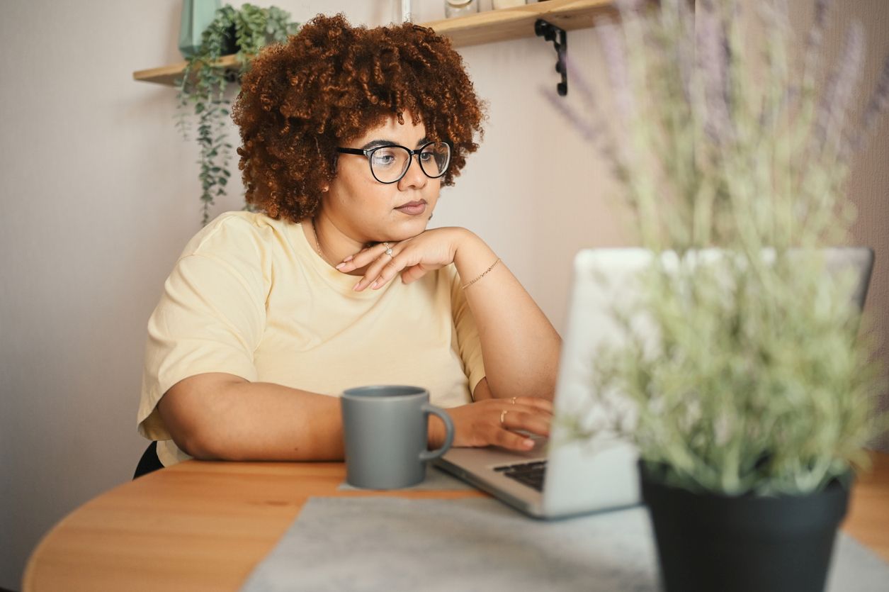 A young woman in glasses looks at her laptop with a coffee cup, surrounded by plants.