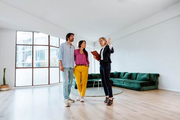 An agent shows a young couple an apartment with large windows and a green sectional couch.