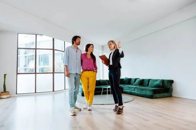 An agent shows a young couple an apartment with large windows and a green sectional couch.
