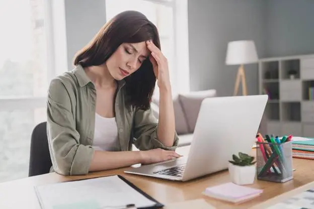 A stressed young woman holds her forehead with her hand as she’s on her laptop.