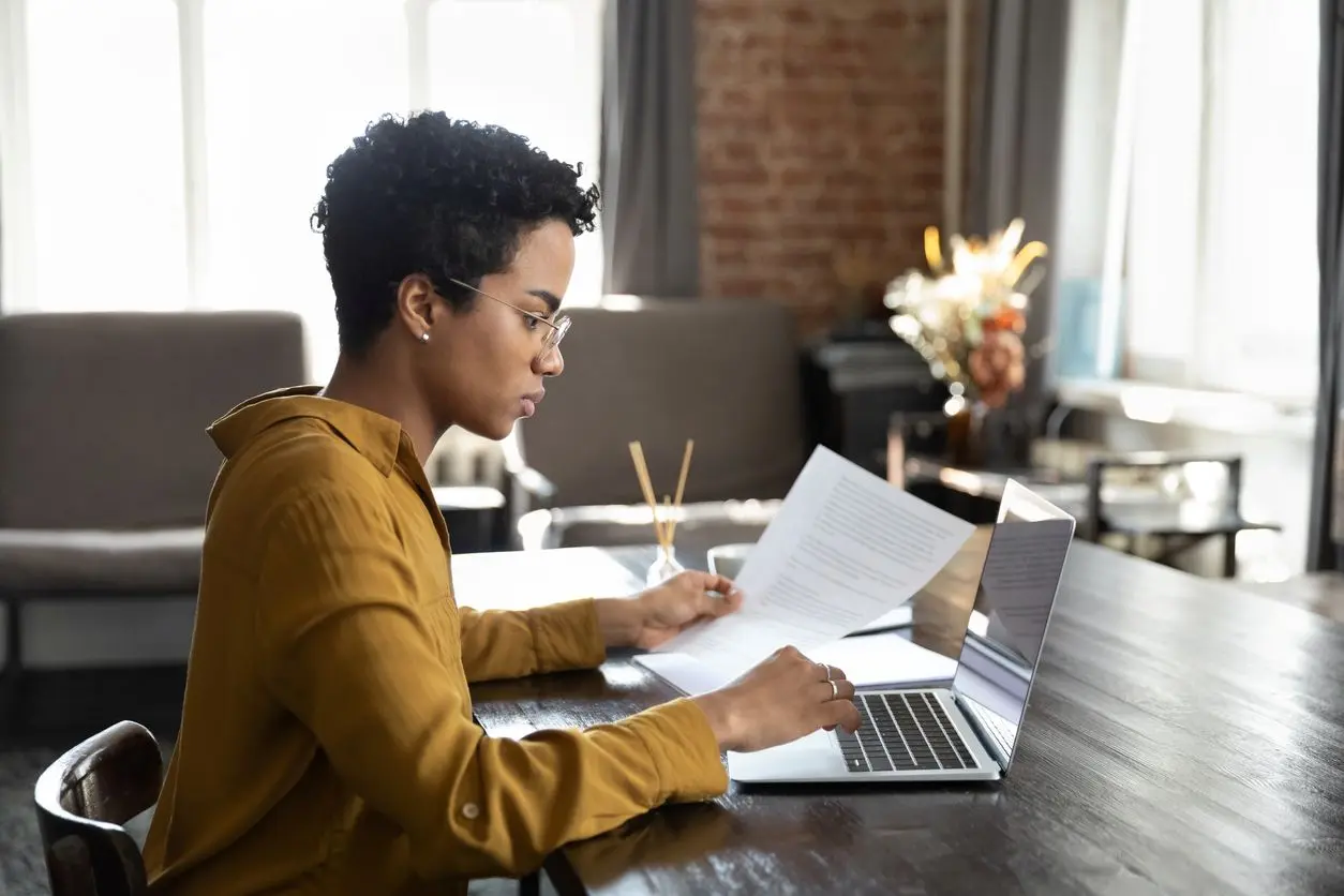 A focused young woman reviews a paper document with a laptop while sitting at a wooden table.