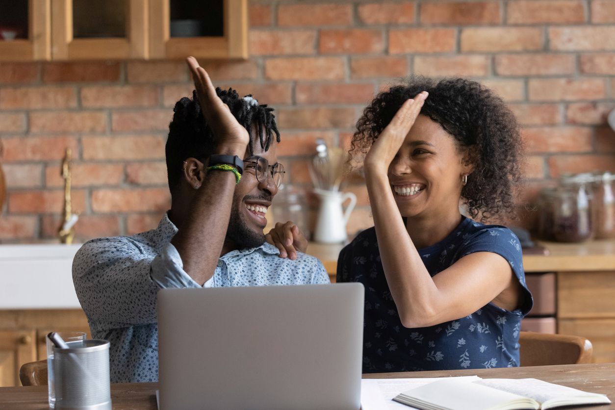 A happy, smiling couple high-five while sitting at a wooden table with a laptop open in front of them.