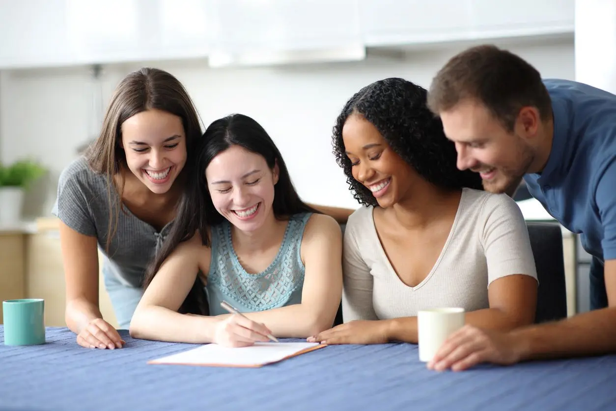 A group of four smiling roommates read over a document on a table with a blue tablecloth