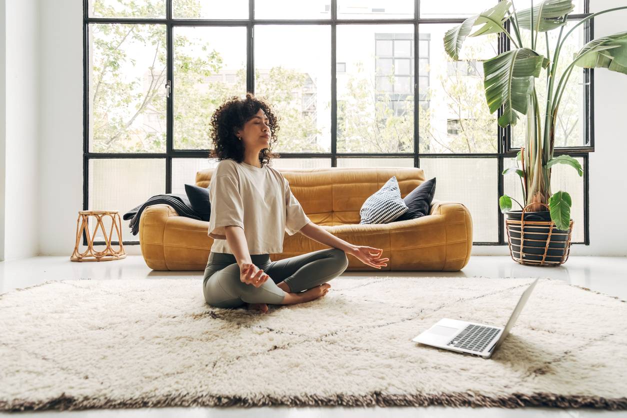 A young woman sits cross-legged on the floor, meditating, in a bright room with an orange couch behind her.