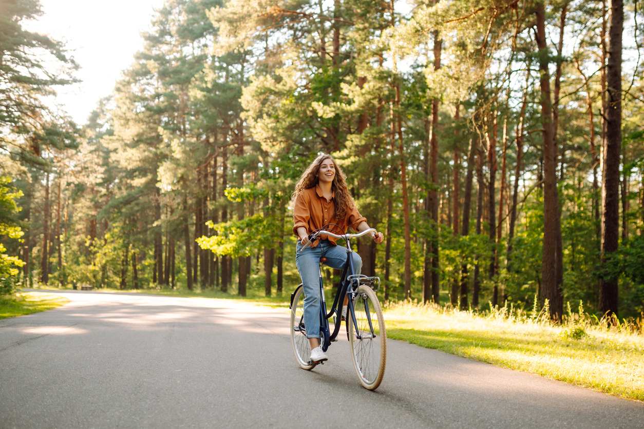 A smiling young woman rides a bike in the middle of a trail surrounded by trees on a sunny day.