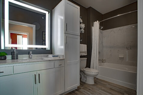 Stylish bathroom at Allure in Centerville, Ohio by Hills Properties featuring white cabinetry, quartz countertops, illuminated mirror, tiled tub surround, and modern finishes.