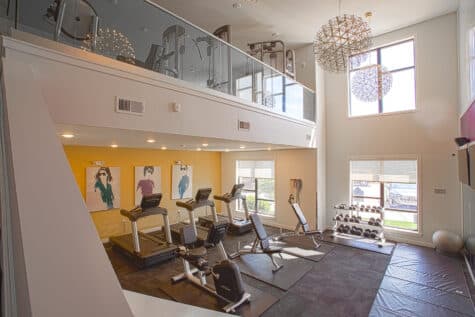 Two-story fitness center at Allure Apartments with cardio equipment, free weights, glass railings, and modern pendant lighting.