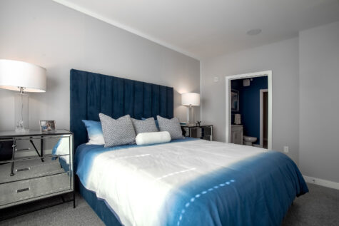 Modern bedroom at Allure in Centerville, Ohio by HILLS Properties with a plush blue accent bed, decorative pillows, mirrored furniture, neutral gray walls, and an en-suite bathroom visible through the open doorway.