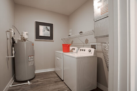 Modern laundry room at Allure in Centerville, Ohio by Hills Properties featuring a white washer and dryer set, wall-mounted wire shelving, wood-look flooring, neutral gray walls, and a utility water heater.