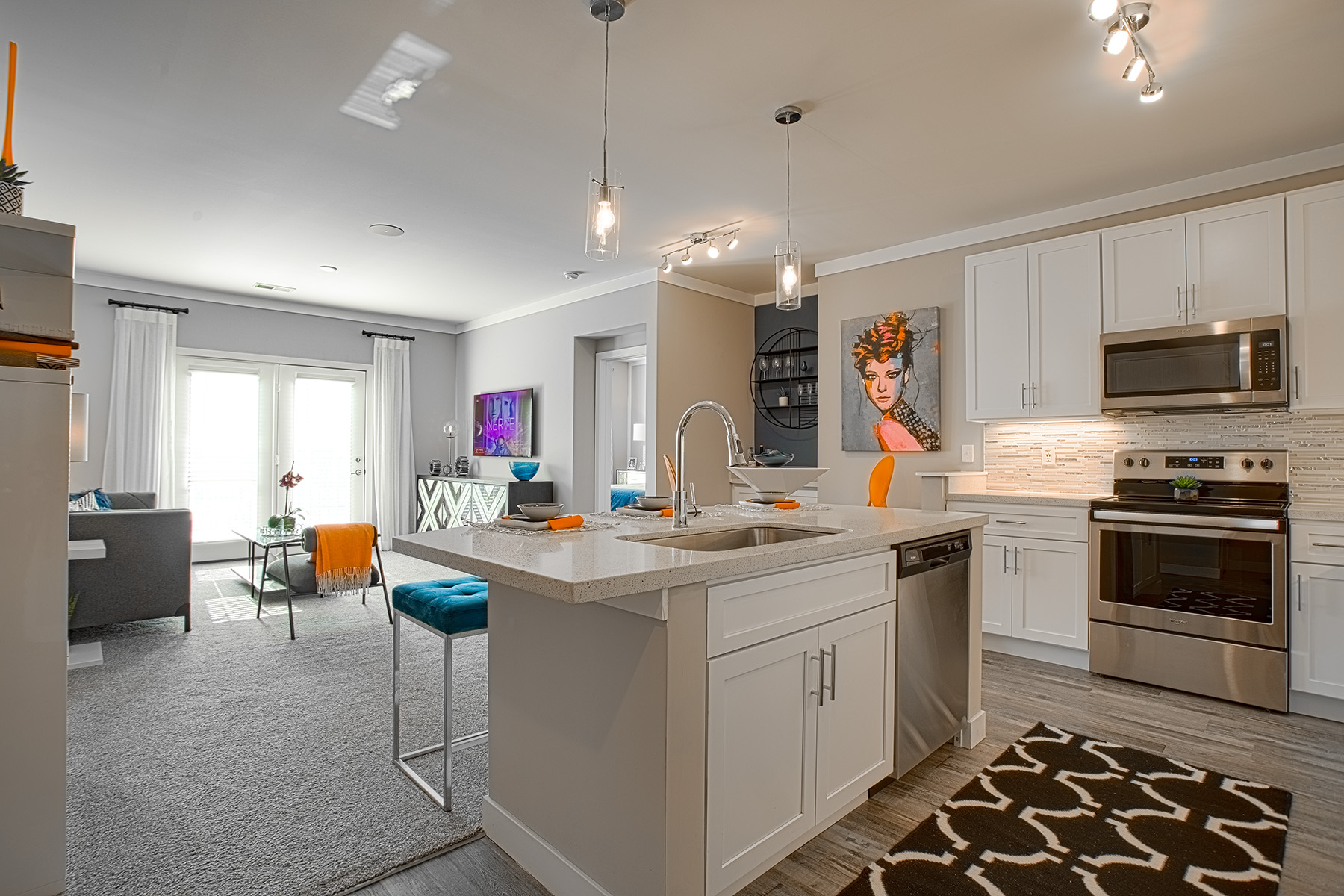 Modern open-concept apartment kitchen and living area at Allure Apartments by HILLS Properties featuring white shaker cabinetry, quartz island countertops, stainless steel appliances, pendant lighting, and colorful contemporary decor accents.