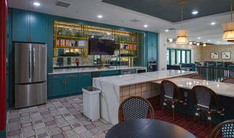 Upscale resident lounge kitchen featuring teal cabinets, patterned tile flooring, gold-accent shelving, oversized quartz island seating, and open access to a contemporary gathering area.