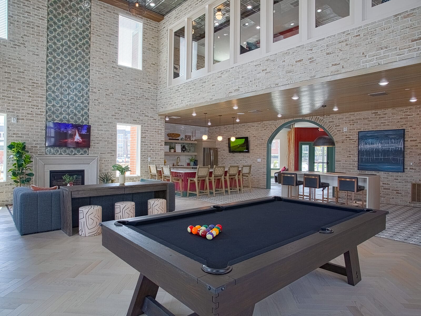 Spacious clubhouse lounge with billiards table, fireplace, open kitchen bar seating, and double-height brick walls with modern industrial décor.