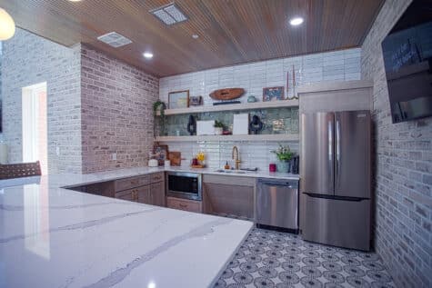 Modern clubhouse kitchenette with quartz countertops, stainless steel appliances, open shelving, patterned tile flooring, and warm wood ceiling accents.