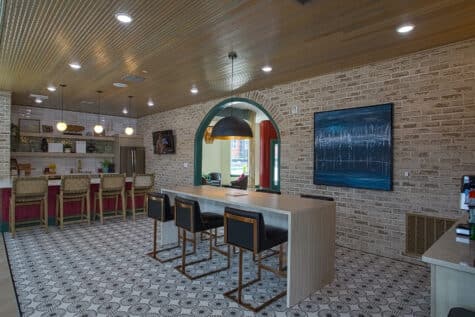 Contemporary resident lounge with communal high-top table, bar seating, patterned tile floors, exposed brick walls, and open entertainment kitchen area.