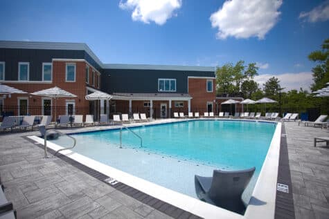 Resort-style swimming pool with in-water lounge seating, shaded umbrellas, and modern clubhouse exterior at Relay 129 by HILLS Properties in Liberty Township, Ohio.