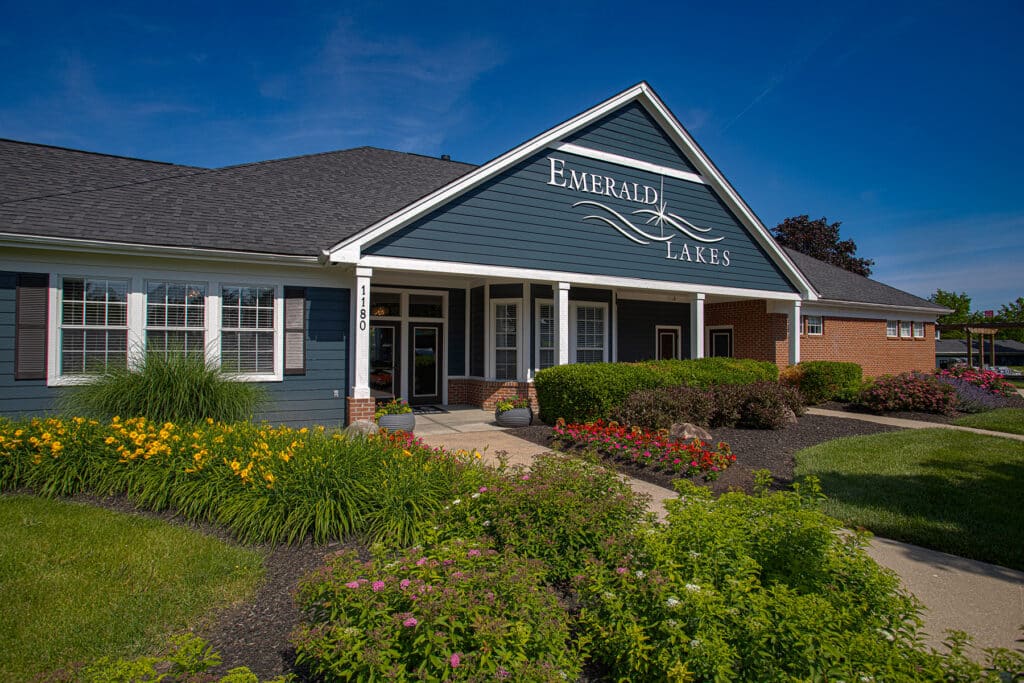 Exterior view of the Emerald Lakes clubhouse in Greenwood, Indiana, showcasing landscaped grounds, colorful flower beds, covered entryway, and modern apartment community architecture by HILLS Properties.