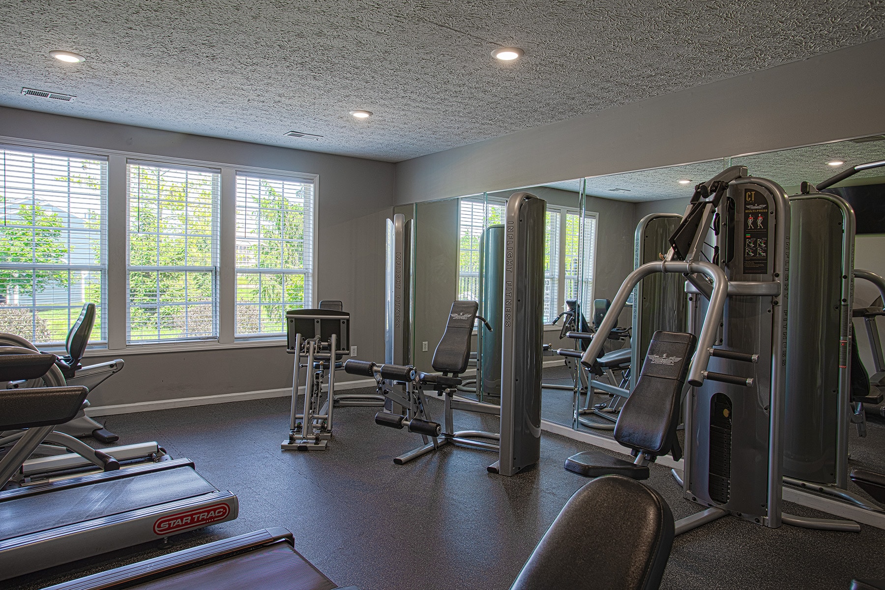 Bright community fitness center at Emerald Lakes in Greenwood, Indiana, with treadmills, weight machines, floor-to-ceiling mirrors, and large windows overlooking the landscaped property by HILLS Properties.