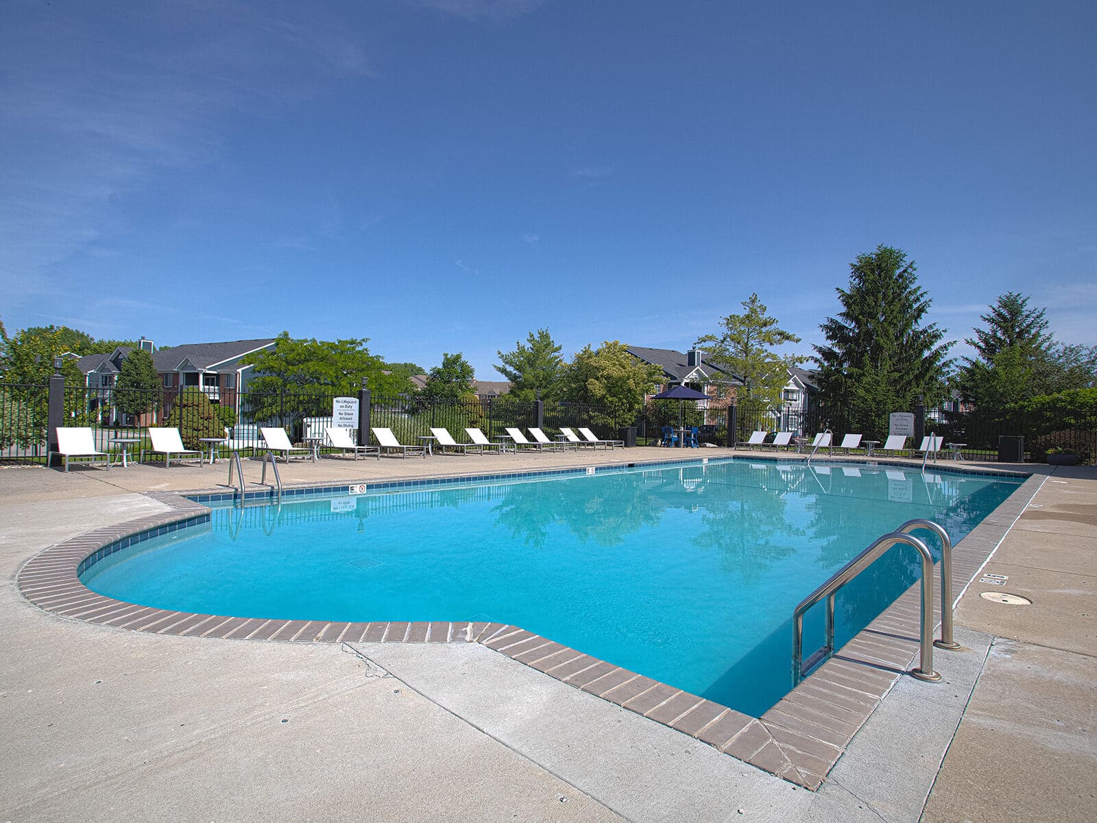 Resort-style outdoor swimming pool with lounge chairs, metal handrails, surrounding landscaping, and apartment community views on a sunny day.