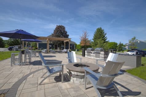 Community outdoor lounge featuring Adirondack chairs around a fire pit, shaded tables with umbrellas, grilling stations, and paved patio space.