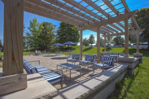 Spacious outdoor patio featuring a large wooden pergola, modern lounge seating, paved gathering area, and open grassy surroundings under a sunny blue sky.