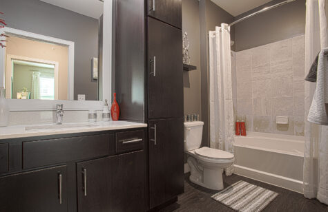 Modern apartment bathroom at Savoy at the Streets of West Chester in West Chester, Ohio featuring dark wood cabinetry, a large vanity mirror, quartz countertop, bathtub with tiled surround, and contemporary finishes with ample storage space.