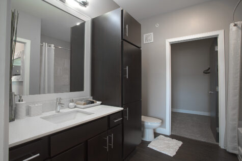 Modern apartment bathroom at Savoy at The Streets of West Chester featuring a white quartz vanity countertop, dark wood cabinetry with abundant storage, a large framed mirror, and a spacious walk-in closet connected to the bathroom. Neutral gray walls and contemporary finishes create a clean, upscale design.