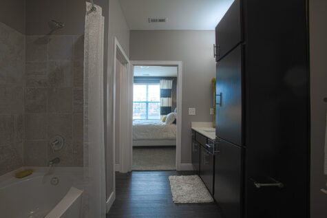 Stylish bathroom at Savoy at The Streets of West Chester showcasing a tub and shower combination with tile surround, dark wood linen cabinets, luxury vinyl plank flooring, and a view into the adjacent bedroom. Modern fixtures and ample storage enhance the functional layout.