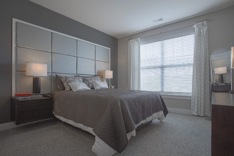 Stylish apartment bedroom at Savoy at the Streets of West Chester in West Chester, Ohio featuring a large upholstered headboard, modern bedside tables with lamps, neutral gray tones, plush carpeting, and a spacious window with natural light.