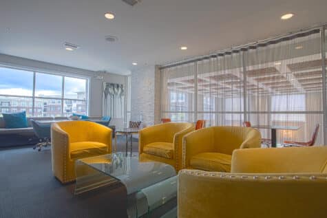 Modern resident lounge at Savoy at The Streets of West Chester in West Chester, Ohio, featuring mustard-yellow lounge chairs, glass coffee tables, floor-to-ceiling windows, and contemporary seating areas by HILLS Properties.