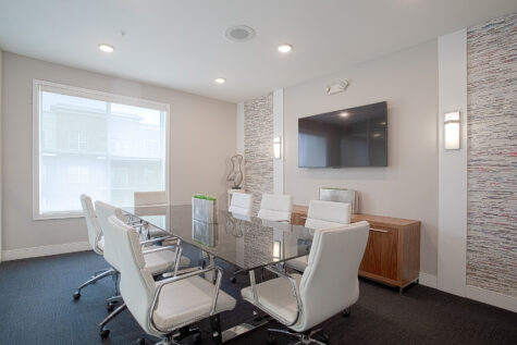Contemporary conference room with a glass-top meeting table surrounded by white leather office chairs. The space features textured wall accents, mounted flat-screen TV, modern lighting, wood cabinetry, and large windows providing natural light.