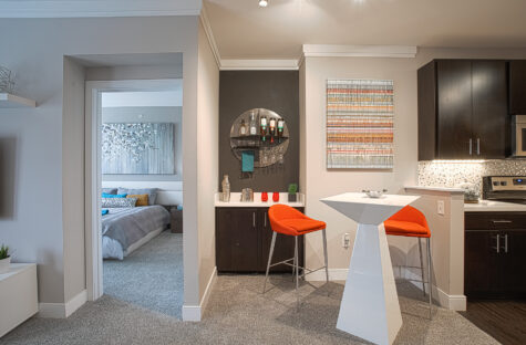 Modern apartment interior at Savoy at the Streets of West Chester in West Chester, Ohio featuring a built-in dry bar with cabinetry and shelving, contemporary dining area with orange bar stools, and an open-concept kitchen with dark cabinets and sleek finishes.