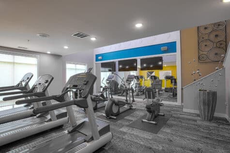 Modern fitness center with treadmills, ellipticals, and stationary bikes arranged on patterned carpet flooring. Large wall mirrors and mounted TVs create a bright, contemporary workout space with recessed lighting and colorful accent walls.